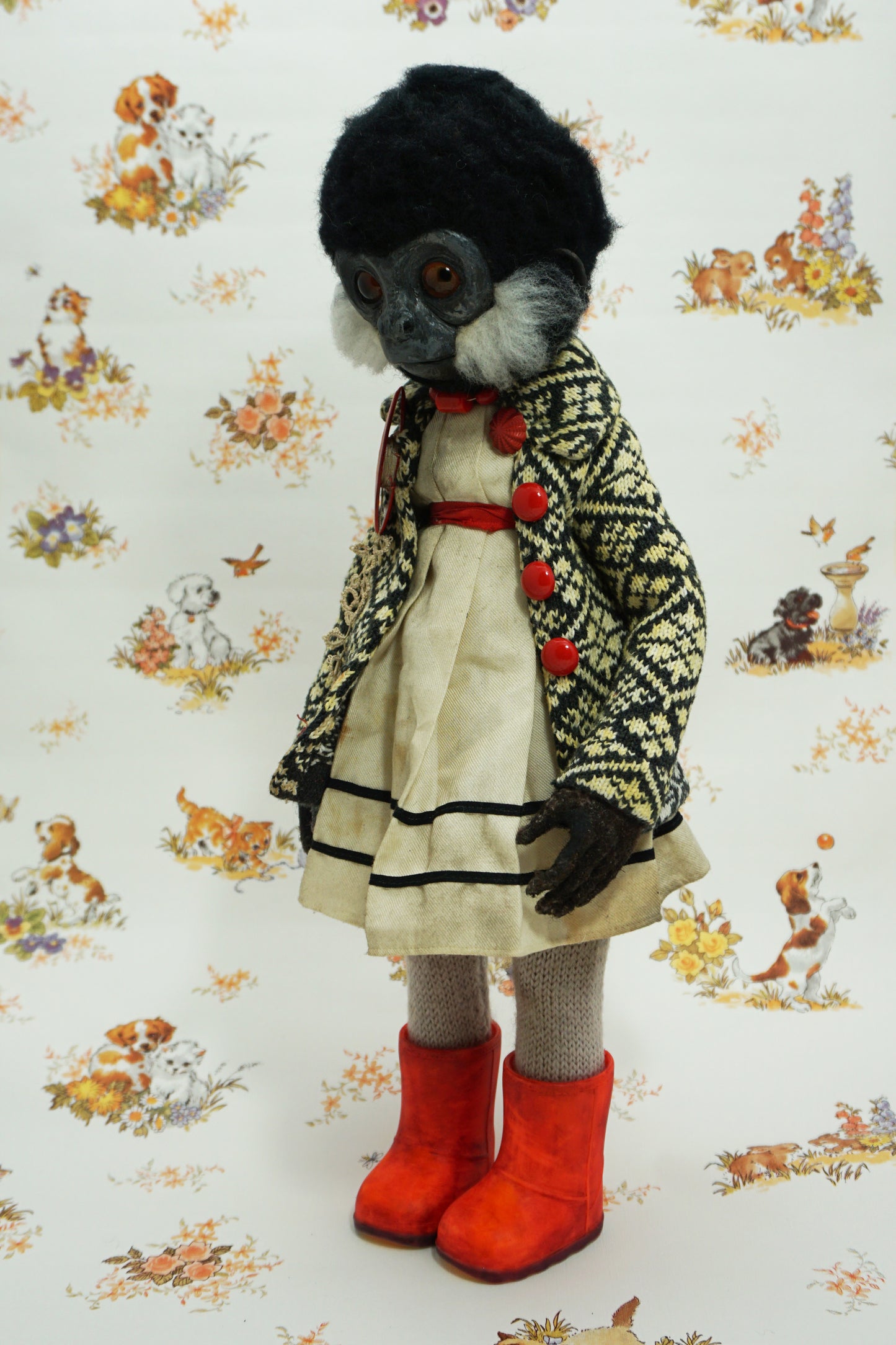 A whimsical doll with a monkey-like face, wearing a patterned coat, white dress, and bright red boots. The background features a vintage-style wallpaper with illustrations of animals and flowers.
