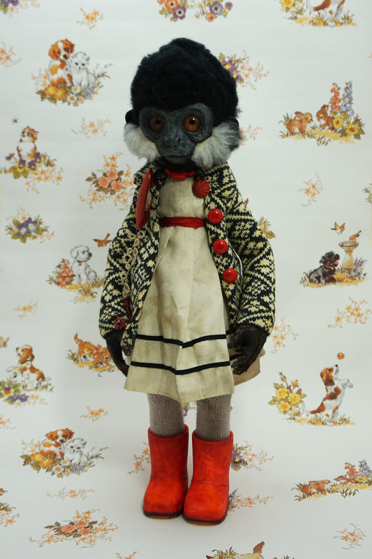 A whimsical doll with a monkey-like face, wearing a patterned coat, white dress, and bright red boots. The background features a vintage-style wallpaper with illustrations of animals and flowers.