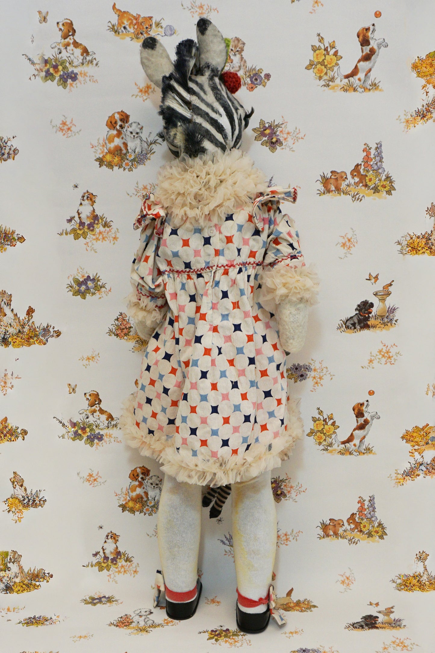 A plush zebra with a patterned dress and red pom-poms stands against a whimsical animal-themed wallpaper. They hold a small zebra toy, and wear black shoes with matching patterned socks.