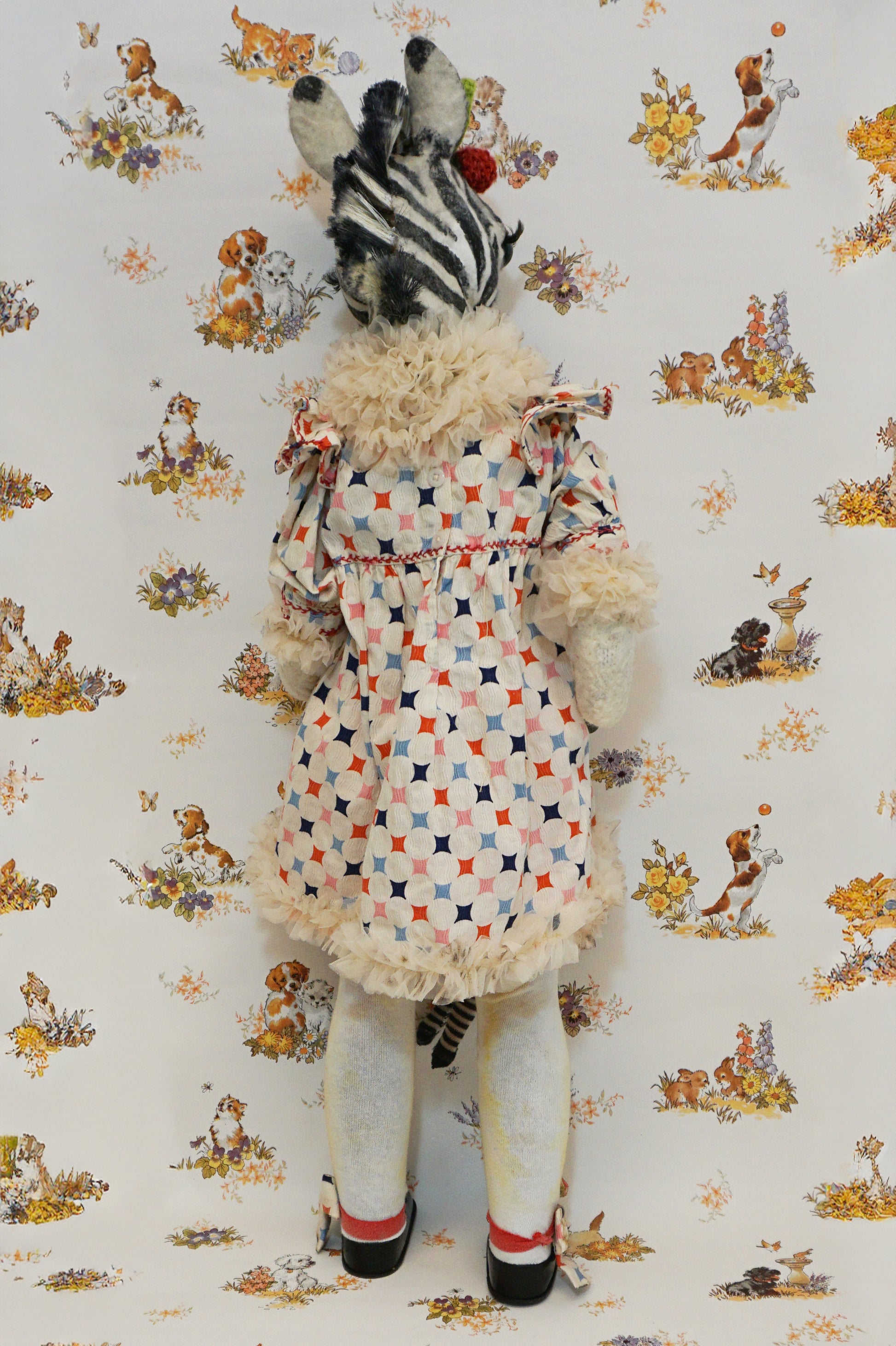 A plush zebra with a patterned dress and red pom-poms stands against a whimsical animal-themed wallpaper. They hold a small zebra toy, and wear black shoes with matching patterned socks.