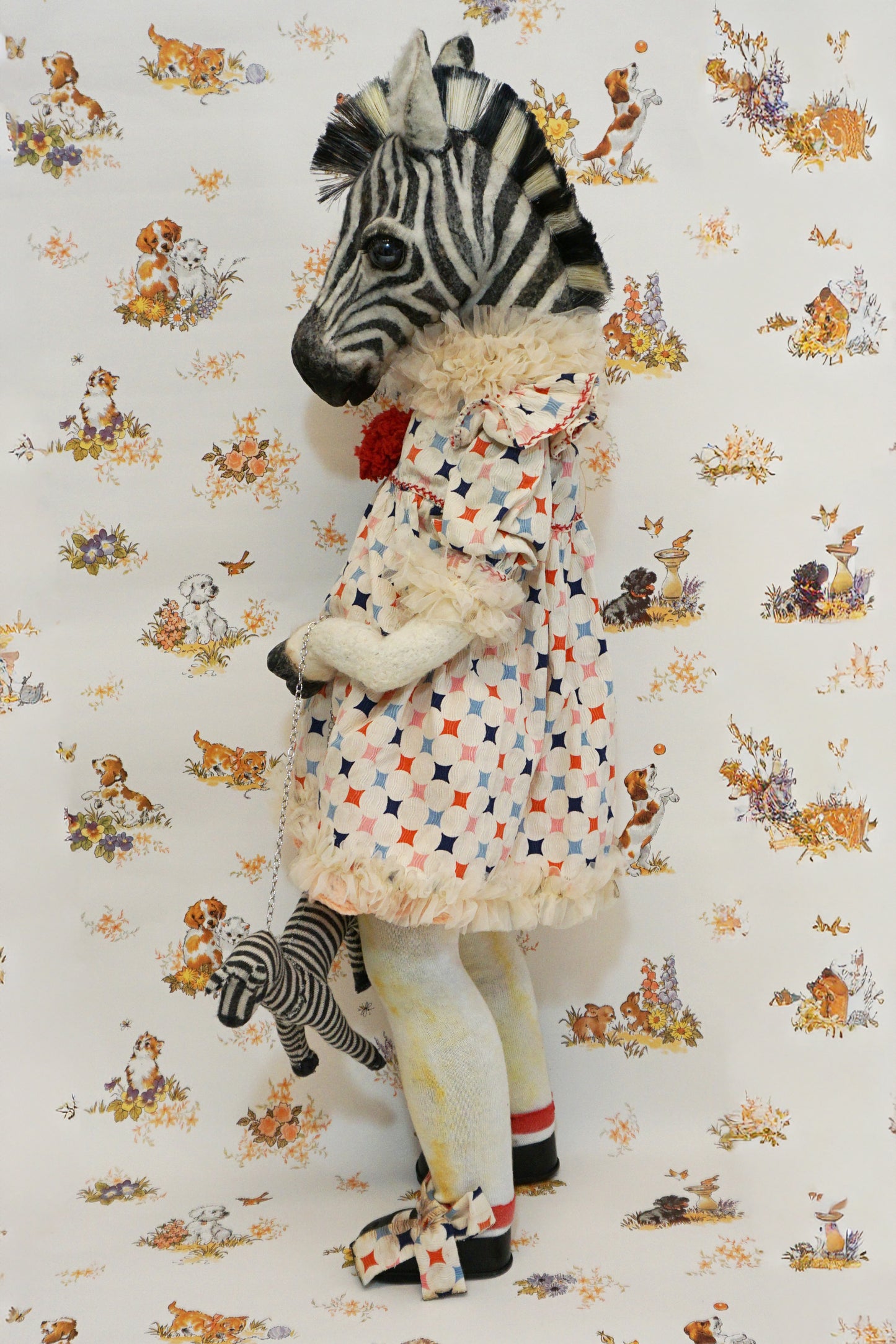 A plush zebra with a patterned dress and red pom-poms stands against a whimsical animal-themed wallpaper. They hold a small zebra toy, and wear black shoes with matching patterned socks.