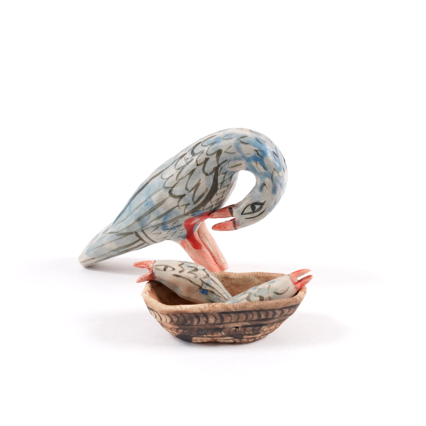 A painted ceramic pelican whistle in shades of blue and grey. There is a ceramic basket with two chicks on the floor next to it all on a plain white background.