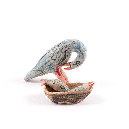 A painted ceramic pelican whistle in shades of blue and grey. There is a ceramic basket with two chicks on the floor next to it all on a plain white background.