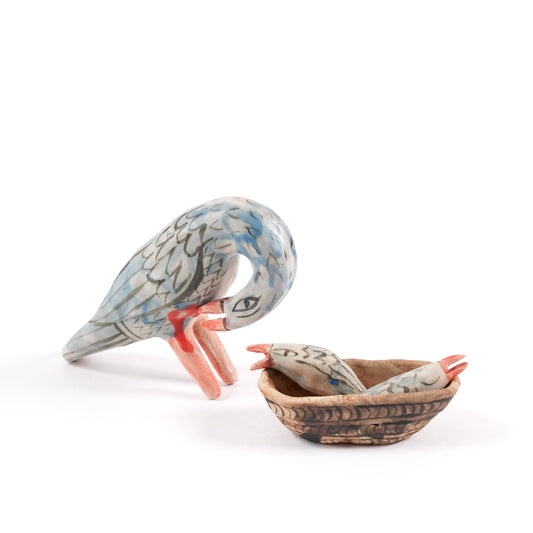 A painted ceramic pelican whistle in shades of blue and grey. There is a ceramic basket with two chicks on the floor next to it all on a plain white background.