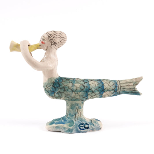 Painted ceramic mermaid whistle blowing a yellow conch shell on a white background, her tail is blue and green.