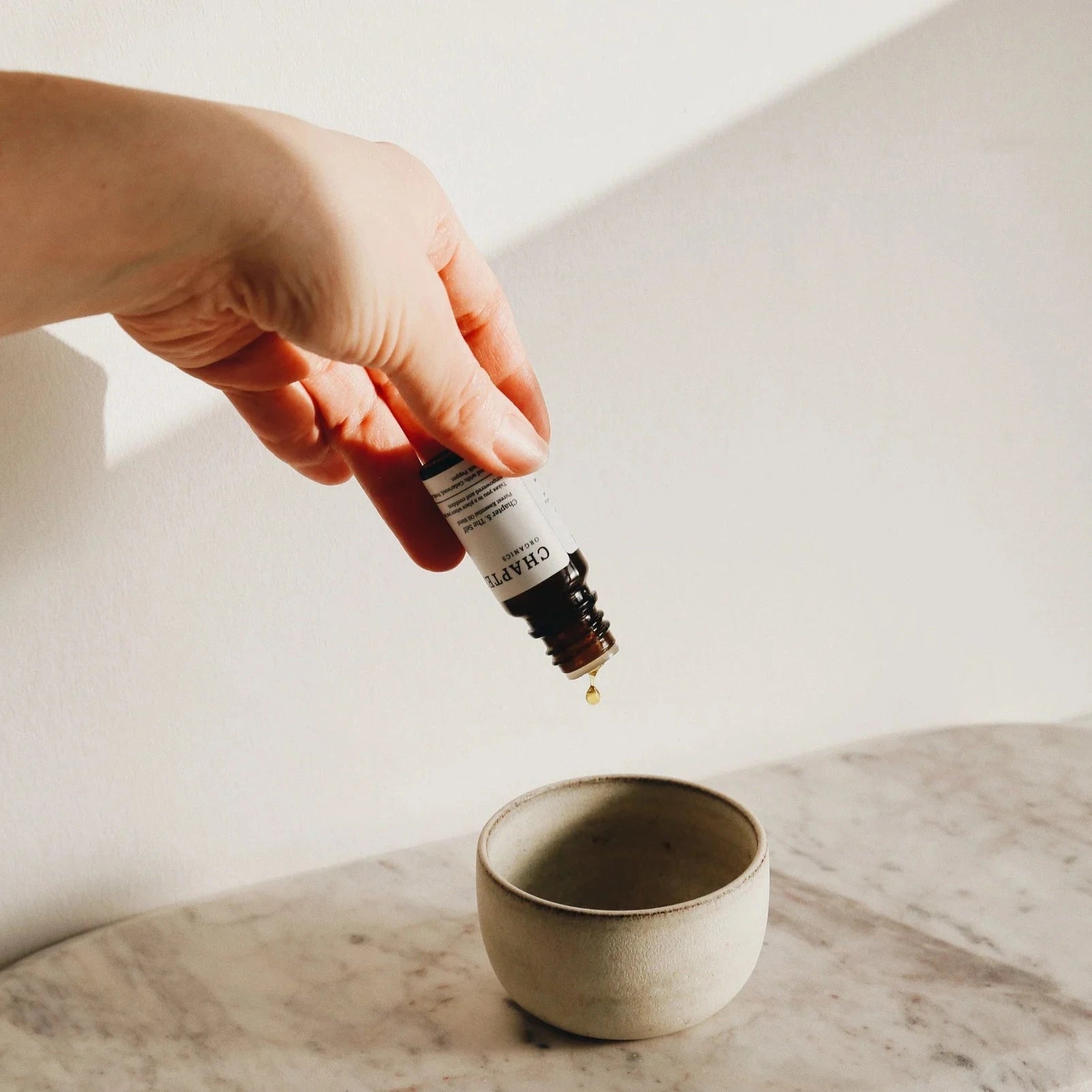 A hand holds a dropper bottle above a small, minimalist ceramic bowl on a marble surface. A single drop of liquid is about to fall into the bowl, with soft natural light illuminating the scene.