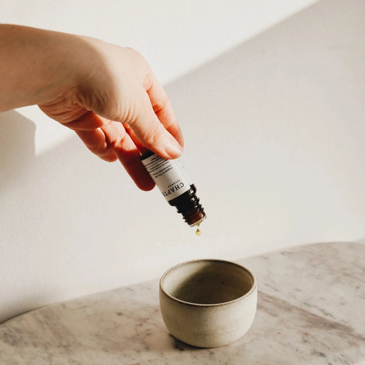 A hand holds a dropper bottle above a small, minimalist ceramic bowl on a marble surface. A single drop of liquid is about to fall into the bowl, with soft natural light illuminating the scene.