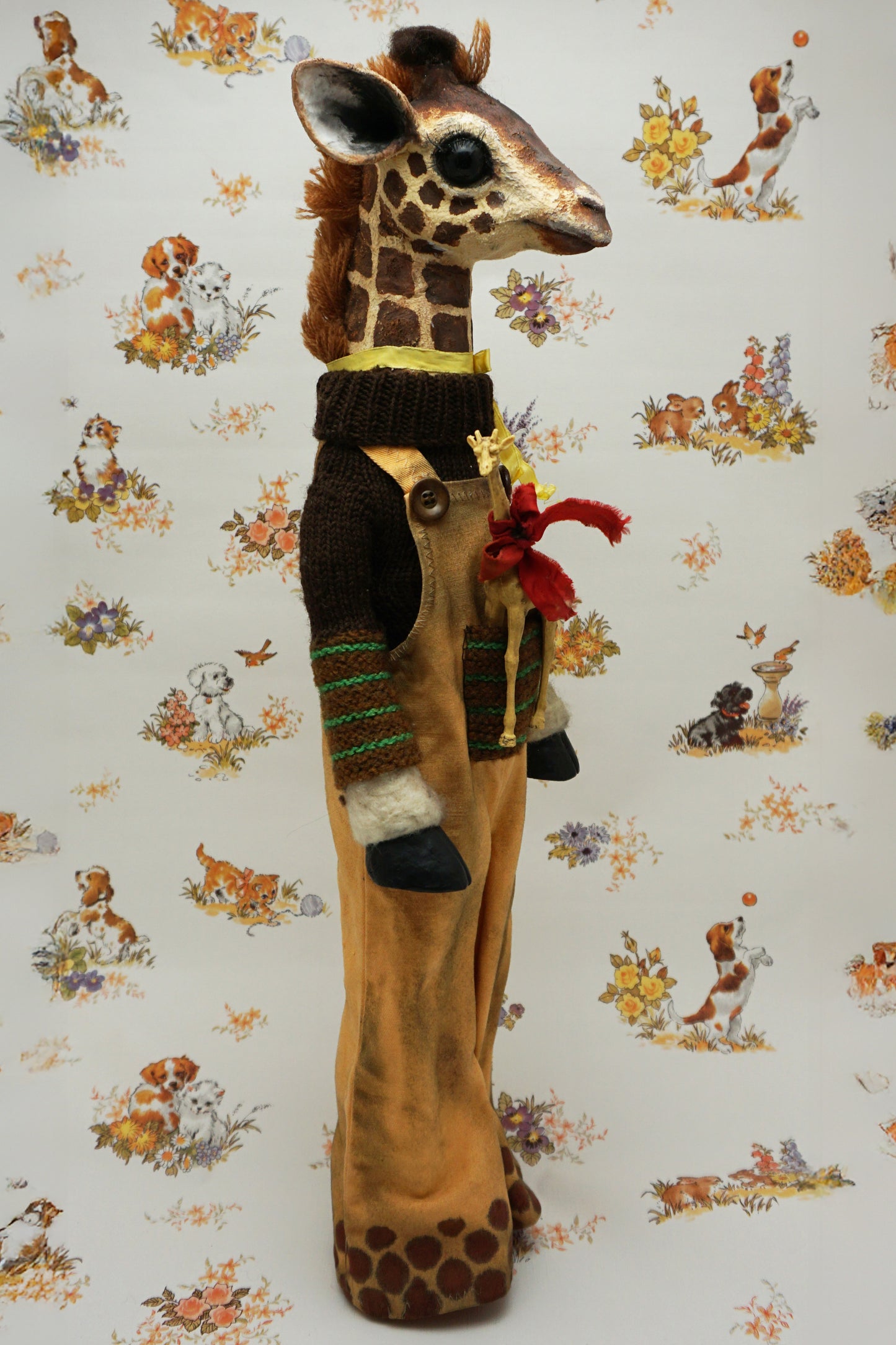 A plush giraffe dressed in a brown jumper and trousers, with a smaller giraffe toy attached to its front. The background features a whimsical pattern of animals and flowers in soft colours.