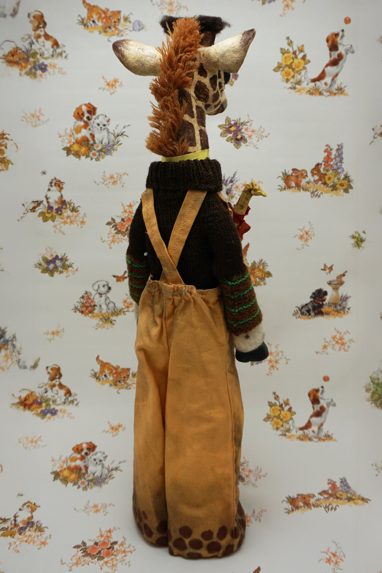 A plush giraffe dressed in a brown jumper and trousers, with a smaller giraffe toy attached to its front. The background features a whimsical pattern of animals and flowers in soft colours.