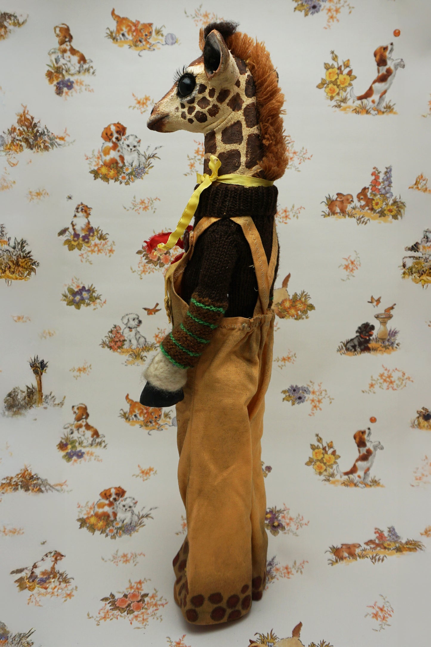 A plush giraffe dressed in a brown jumper and trousers, with a smaller giraffe toy attached to its front. The background features a whimsical pattern of animals and flowers in soft colours.