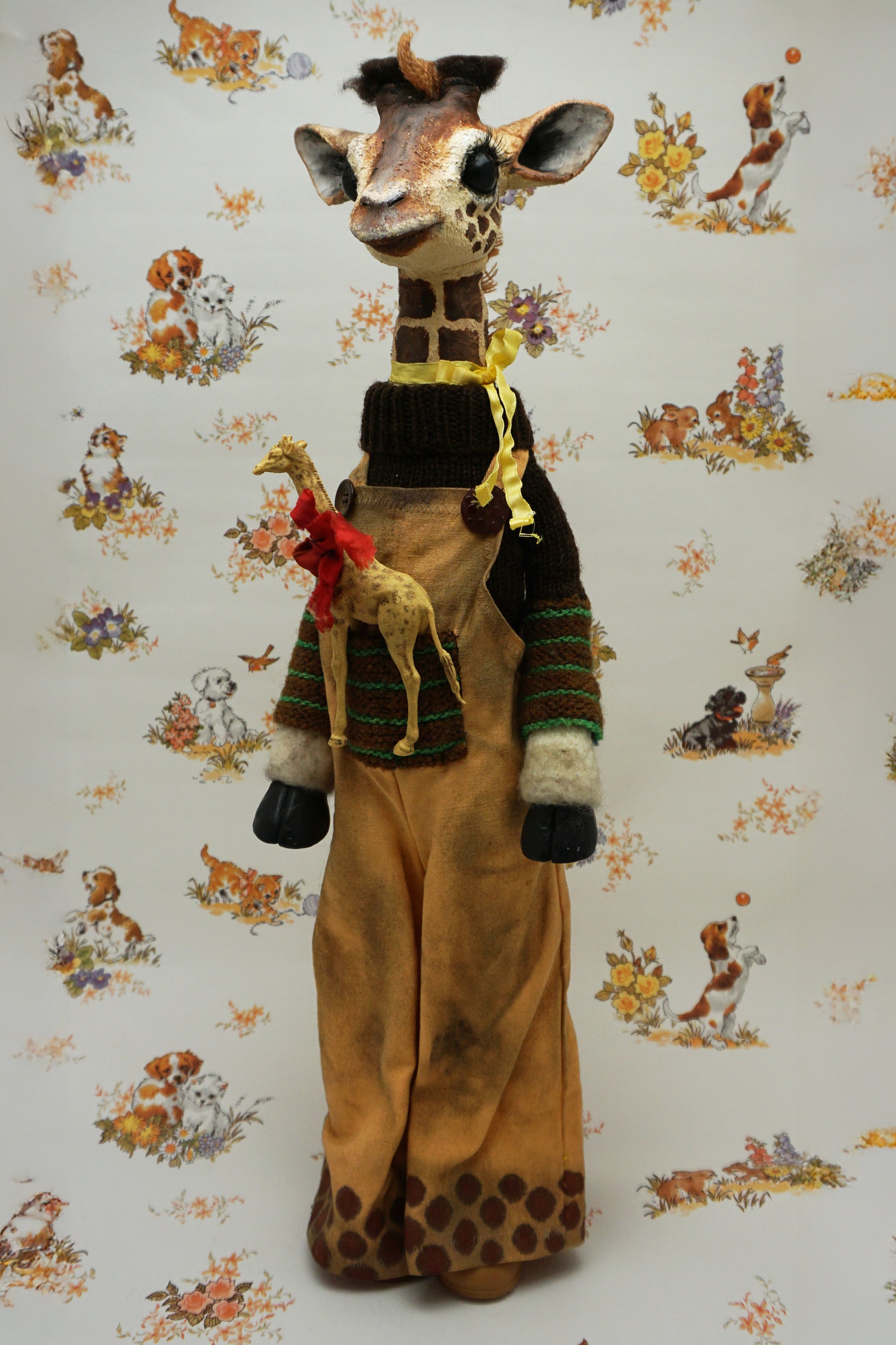 A plush giraffe dressed in a brown jumper and trousers, with a smaller giraffe toy attached to its front. The background features a whimsical pattern of animals and flowers in soft colours.