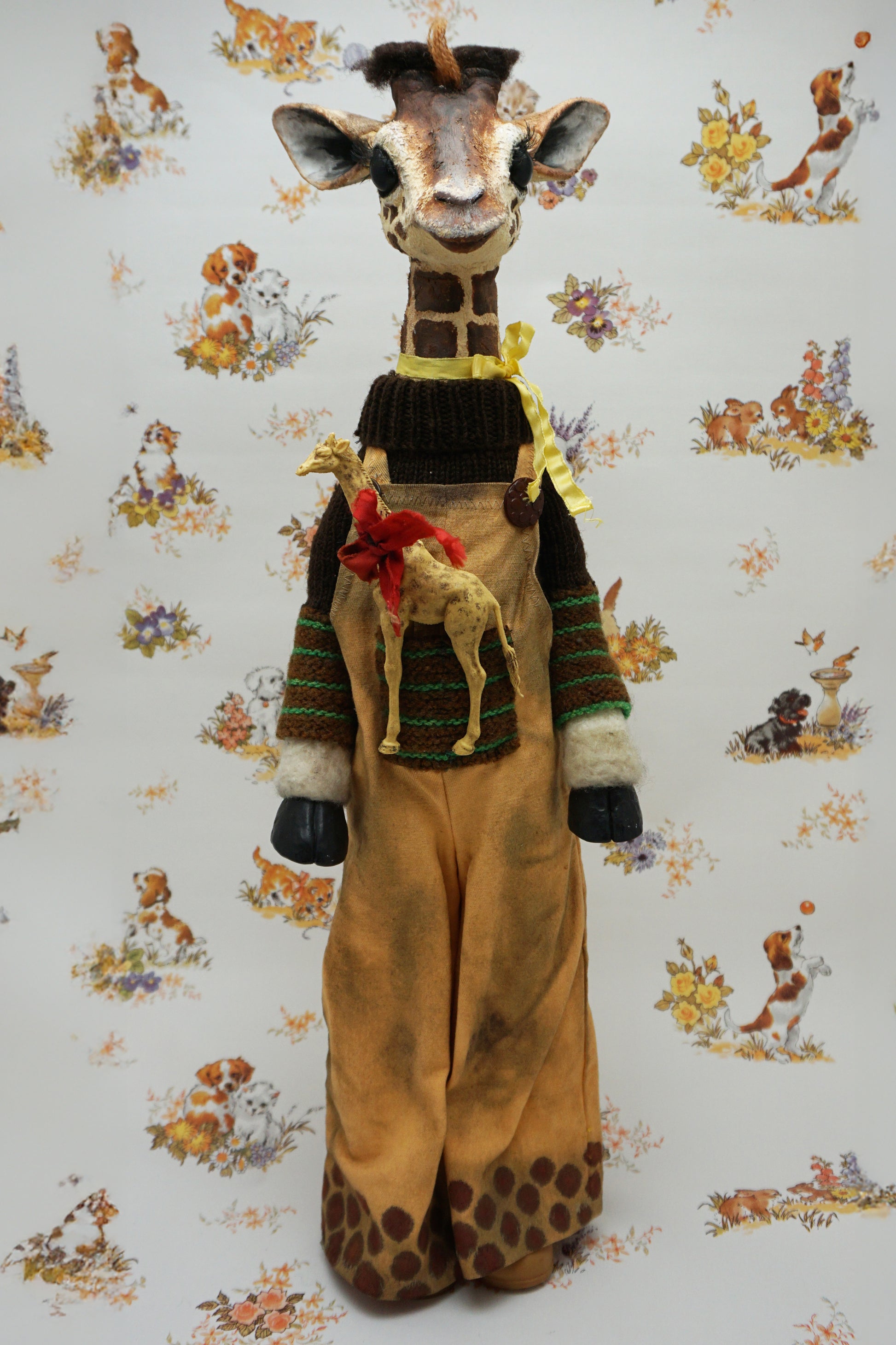 A plush giraffe dressed in a brown jumper and trousers, with a smaller giraffe toy attached to its front. The background features a whimsical pattern of animals and flowers in soft colours.