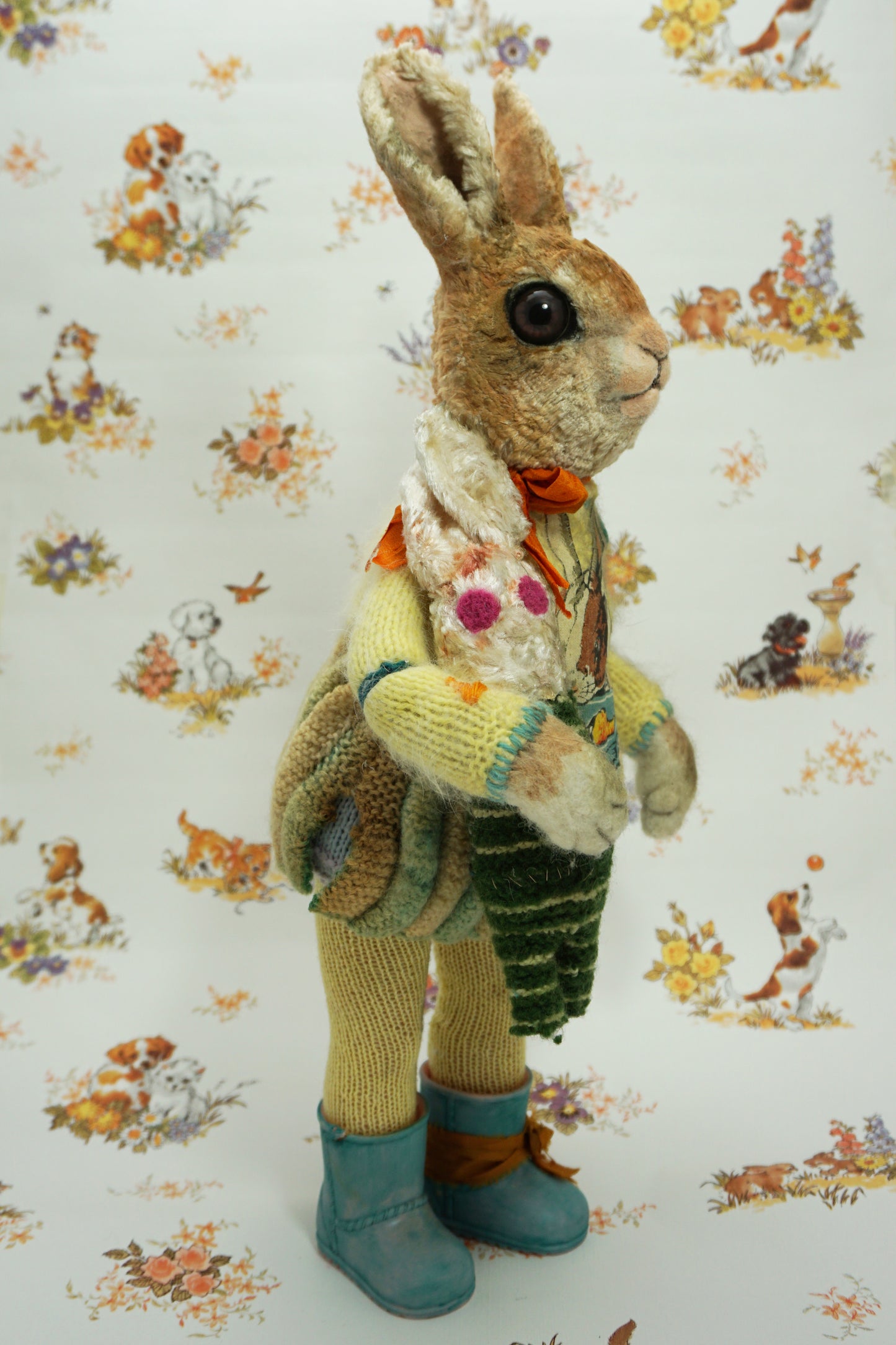 A plush rabbit doll stands upright, wearing a colourful knitted outfit with a rabbit motif. It holds a striped scarf and a small rabbit toy. The background features a whimsical pattern of animals and flowers. The rabbit wears blue boots with orange ribbon the right foot.