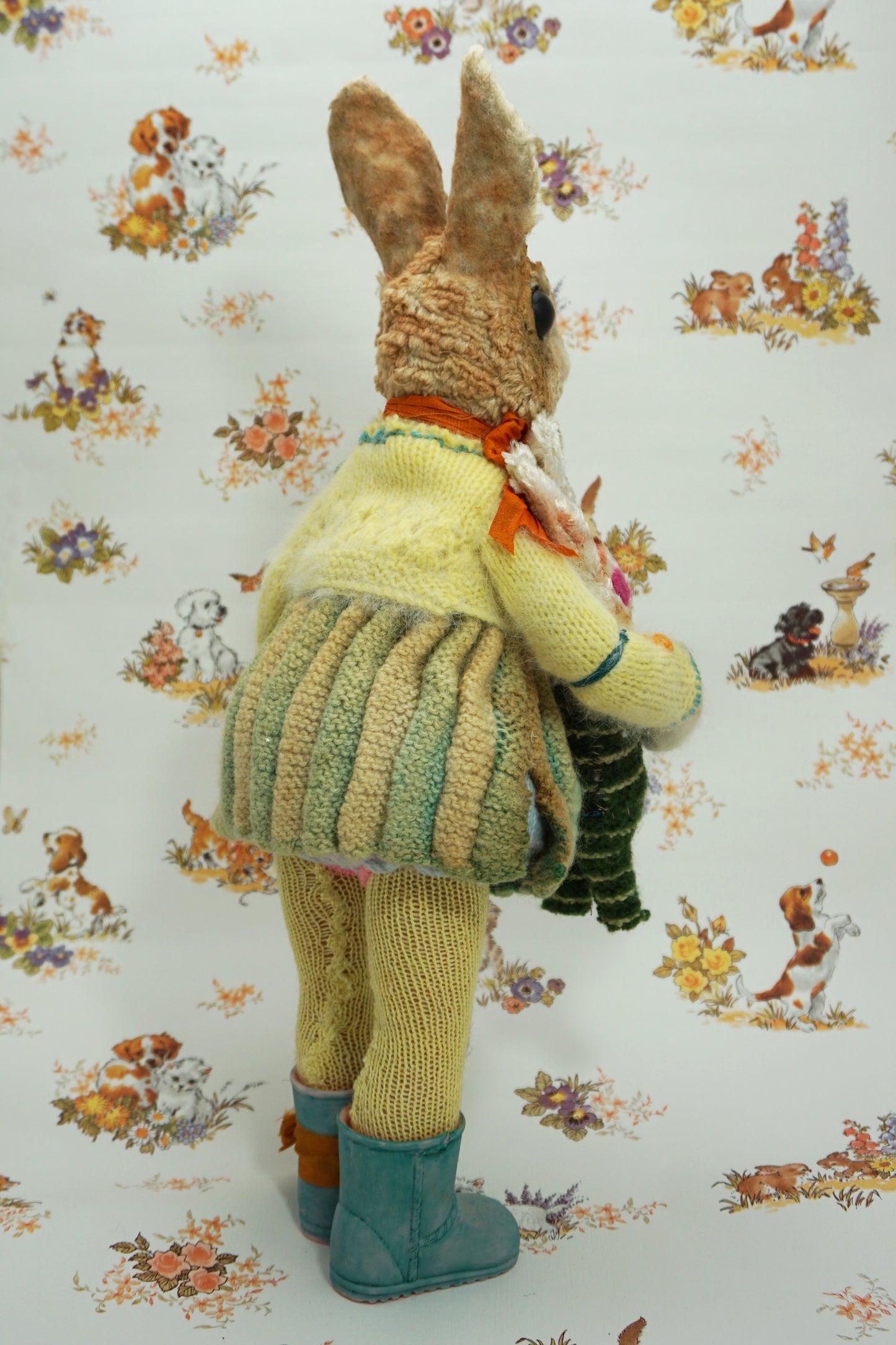A plush rabbit doll stands upright, wearing a colourful knitted outfit with a rabbit motif. It holds a striped scarf and a small rabbit toy. The background features a whimsical pattern of animals and flowers. The rabbit wears blue boots with orange ribbon the right foot.