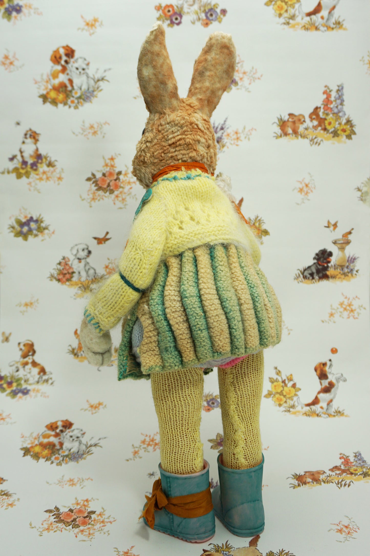 A plush rabbit doll stands upright, wearing a colourful knitted outfit with a rabbit motif. It holds a striped scarf and a small rabbit toy. The background features a whimsical pattern of animals and flowers. The rabbit wears blue boots with orange ribbon the right foot.
