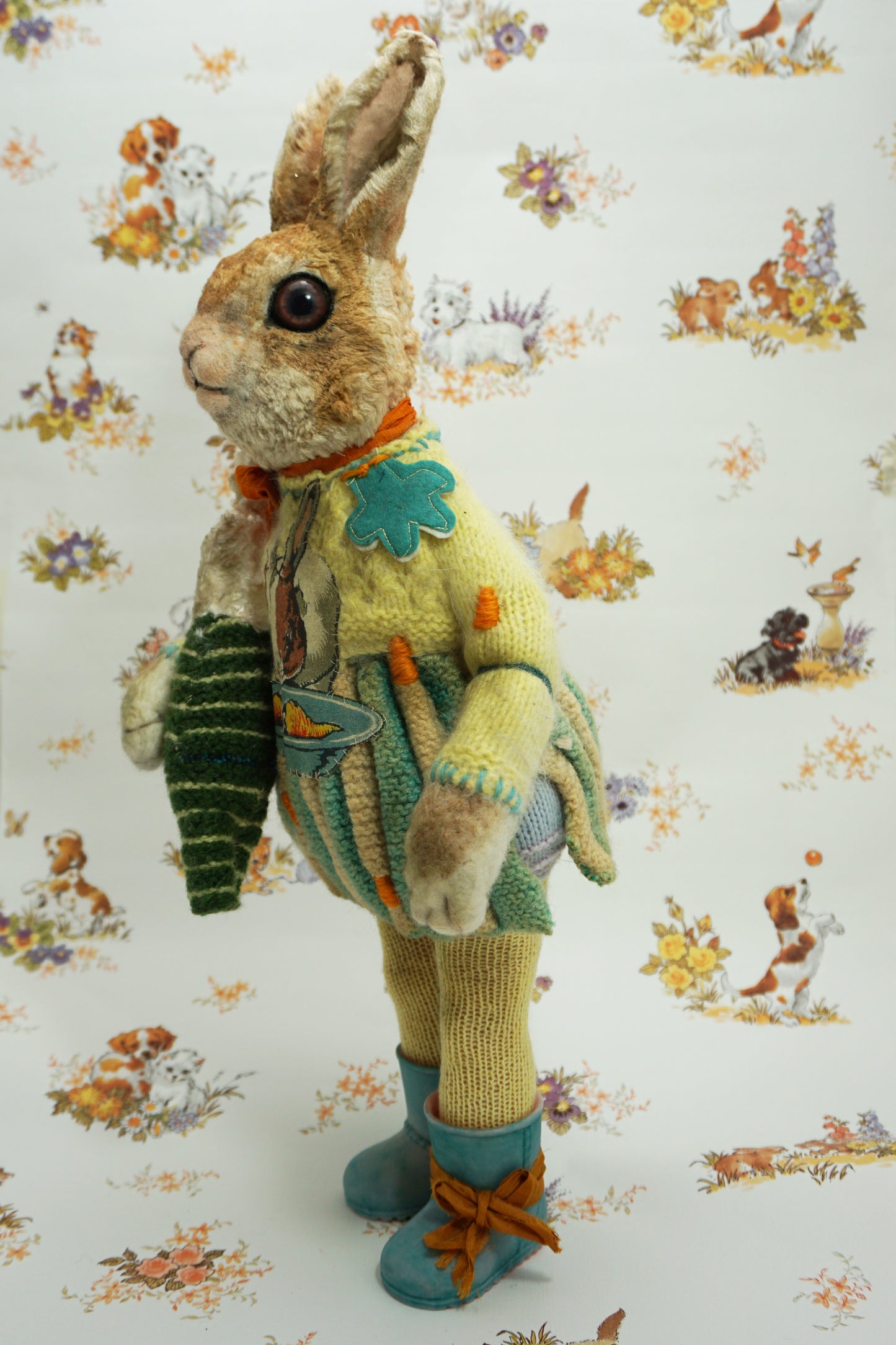 A plush rabbit doll stands upright, wearing a colourful knitted outfit with a rabbit motif. It holds a striped scarf and a small rabbit toy. The background features a whimsical pattern of animals and flowers. The rabbit wears blue boots with orange ribbon the right foot.