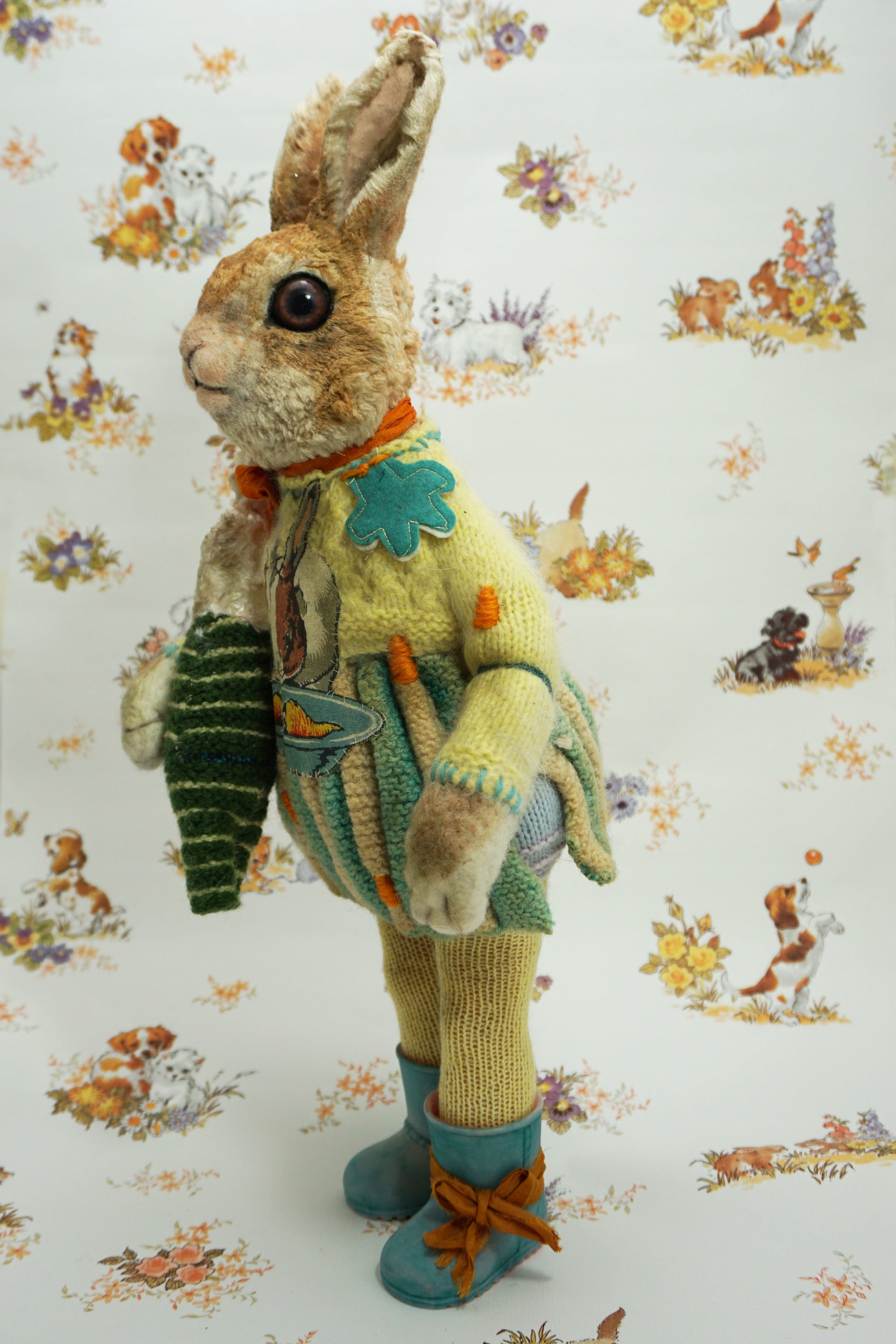 A plush rabbit doll stands upright, wearing a colourful knitted outfit with a rabbit motif. It holds a striped scarf and a small rabbit toy. The background features a whimsical pattern of animals and flowers. The rabbit wears blue boots with orange ribbon the right foot.