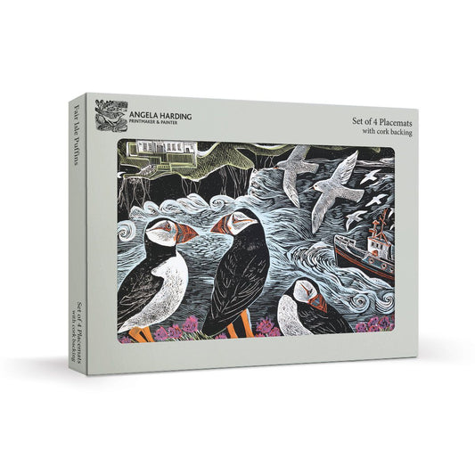 A boxed placemat set titled "Set of 4 Placemats with cork backing" by Angela Harding, shown at a slight angle on a light grey background. The front of the box features a detailed illustration of puffins, seals, waves, and a boat in black, white, grey, and small orange and pink accents.