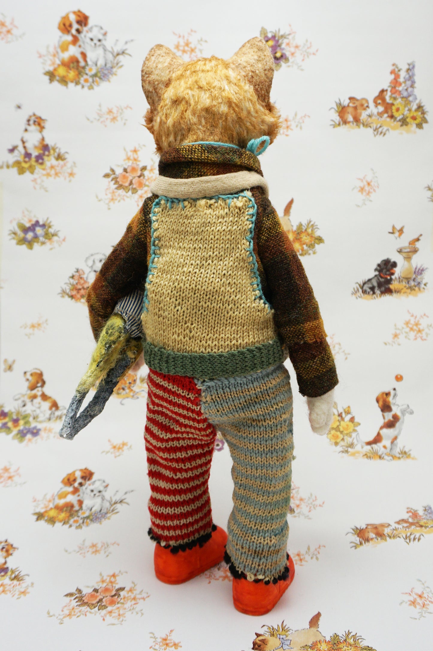 A whimsical cat doll stands upright, wearing a colourful knitted outfit with mismatched patterns and bright orange shoes. It holds a small yellow toy cat. The background features a playful animal-themed pattern.