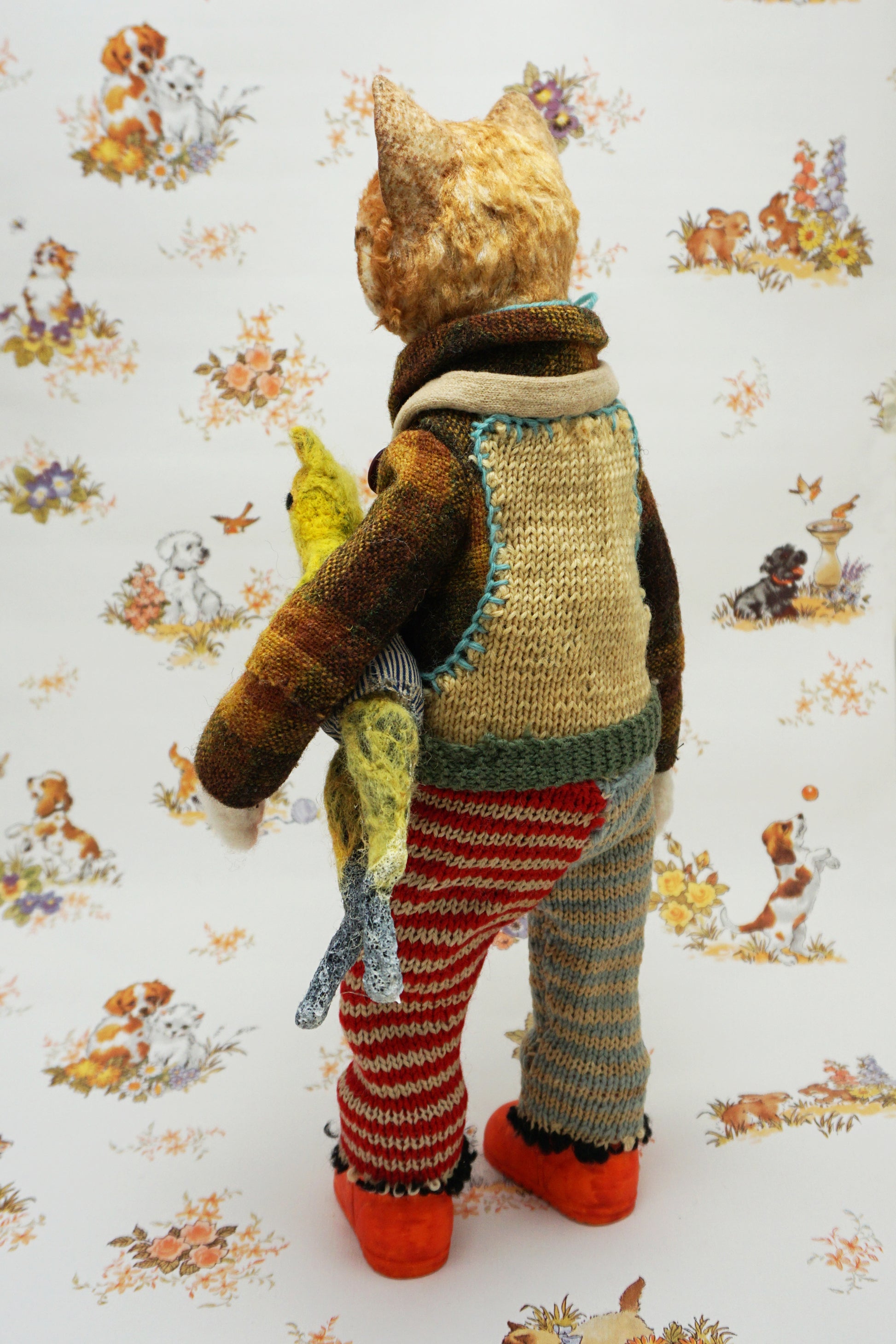 A whimsical cat doll stands upright, wearing a colourful knitted outfit with mismatched patterns and bright orange shoes. It holds a small yellow toy cat. The background features a playful animal-themed pattern.
