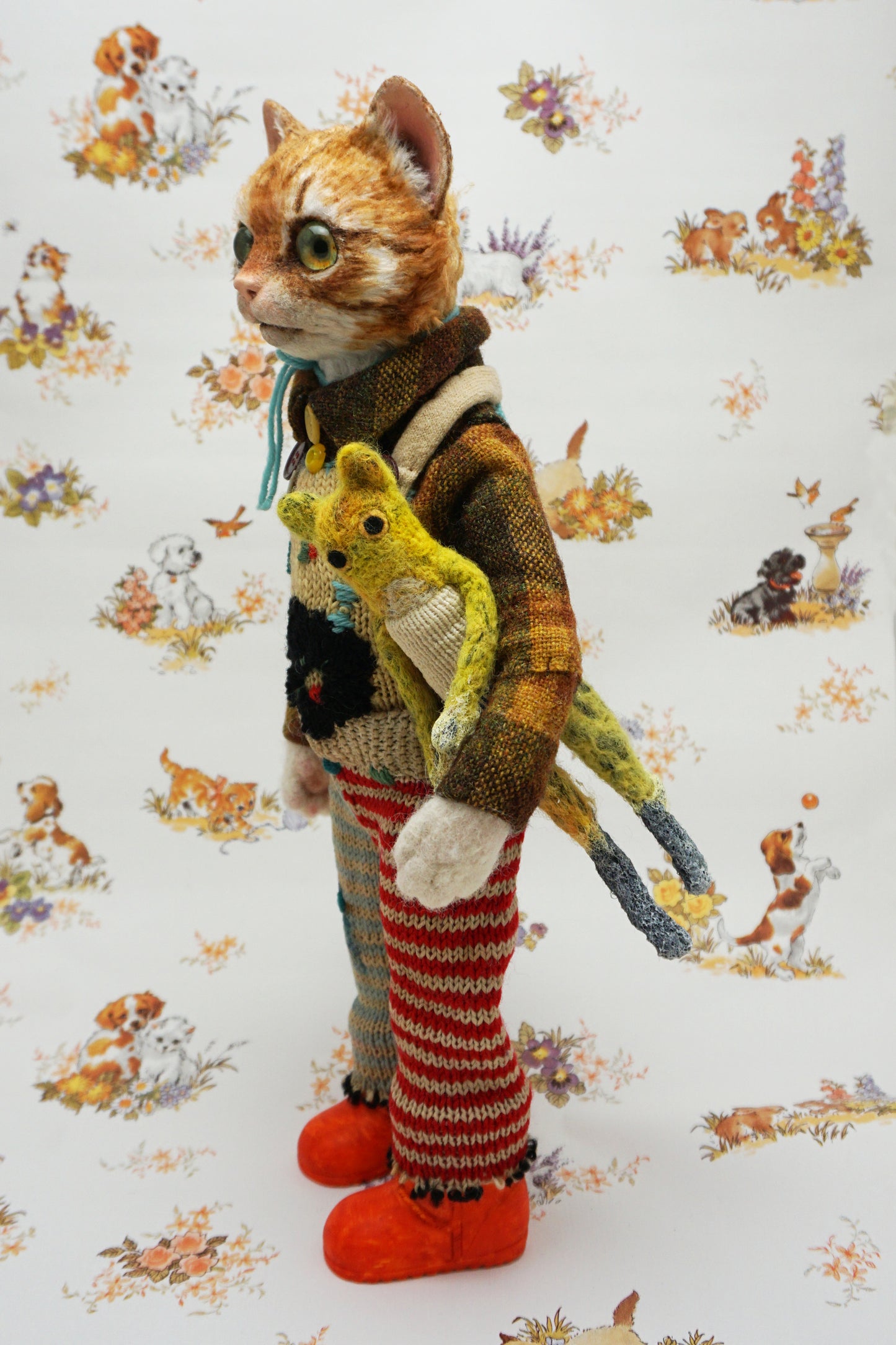 A whimsical cat doll stands upright, wearing a colourful knitted outfit with mismatched patterns and bright orange shoes. It holds a small yellow toy cat. The background features a playful animal-themed pattern.