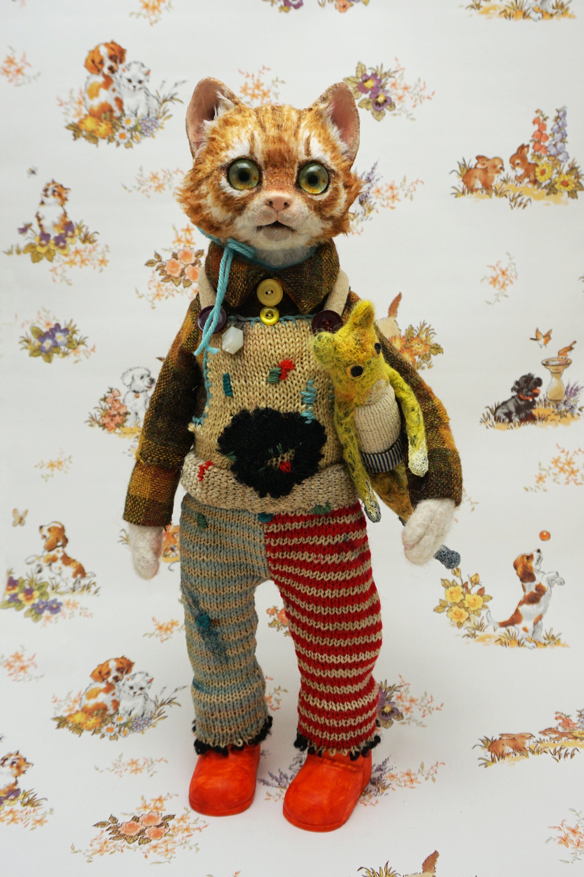 A whimsical cat doll stands upright, wearing a colourful knitted outfit with mismatched patterns and bright orange shoes. It holds a small yellow toy cat. The background features a playful animal-themed pattern.