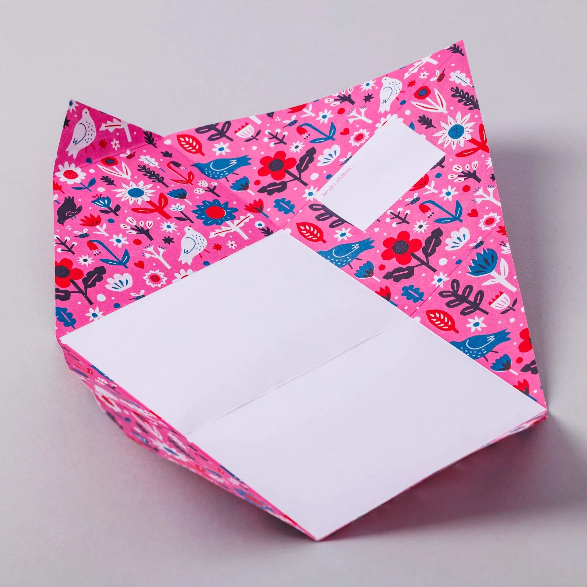 A vibrant pink origami envelope featuring a floral and bird pattern. The envelope is partially open, revealing a blank white section for writing. The background is a neutral grey, highlighting the colourful design of the envelope.