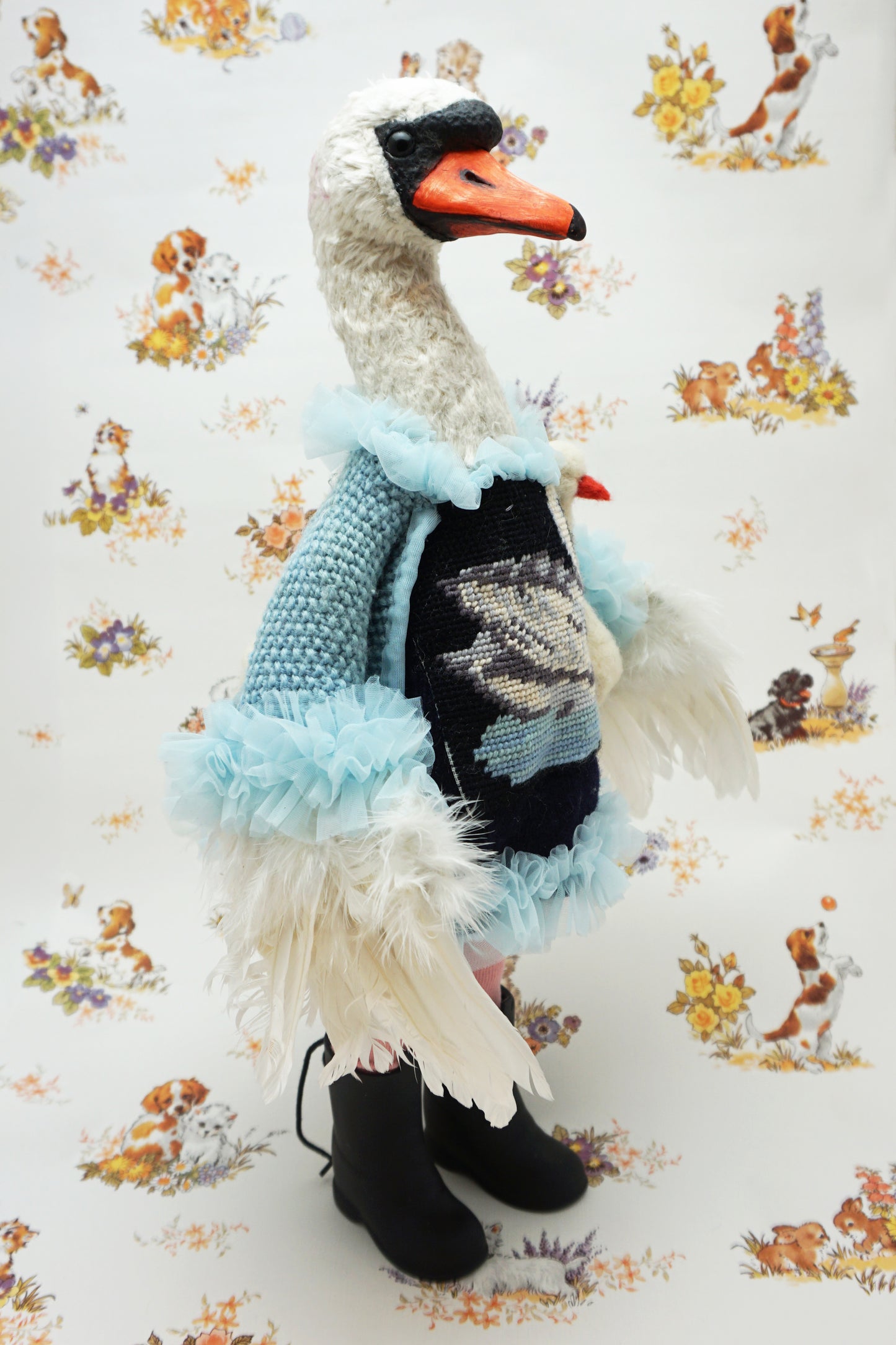 A plush swan holding a smaller swan toy dressed in a blue knitted jumper with a swan design, pink leggings, and black boots. It stands against a backdrop featuring illustrations of dogs and flowers. The swan's jumper has a fluffy blue collar and cuffs.