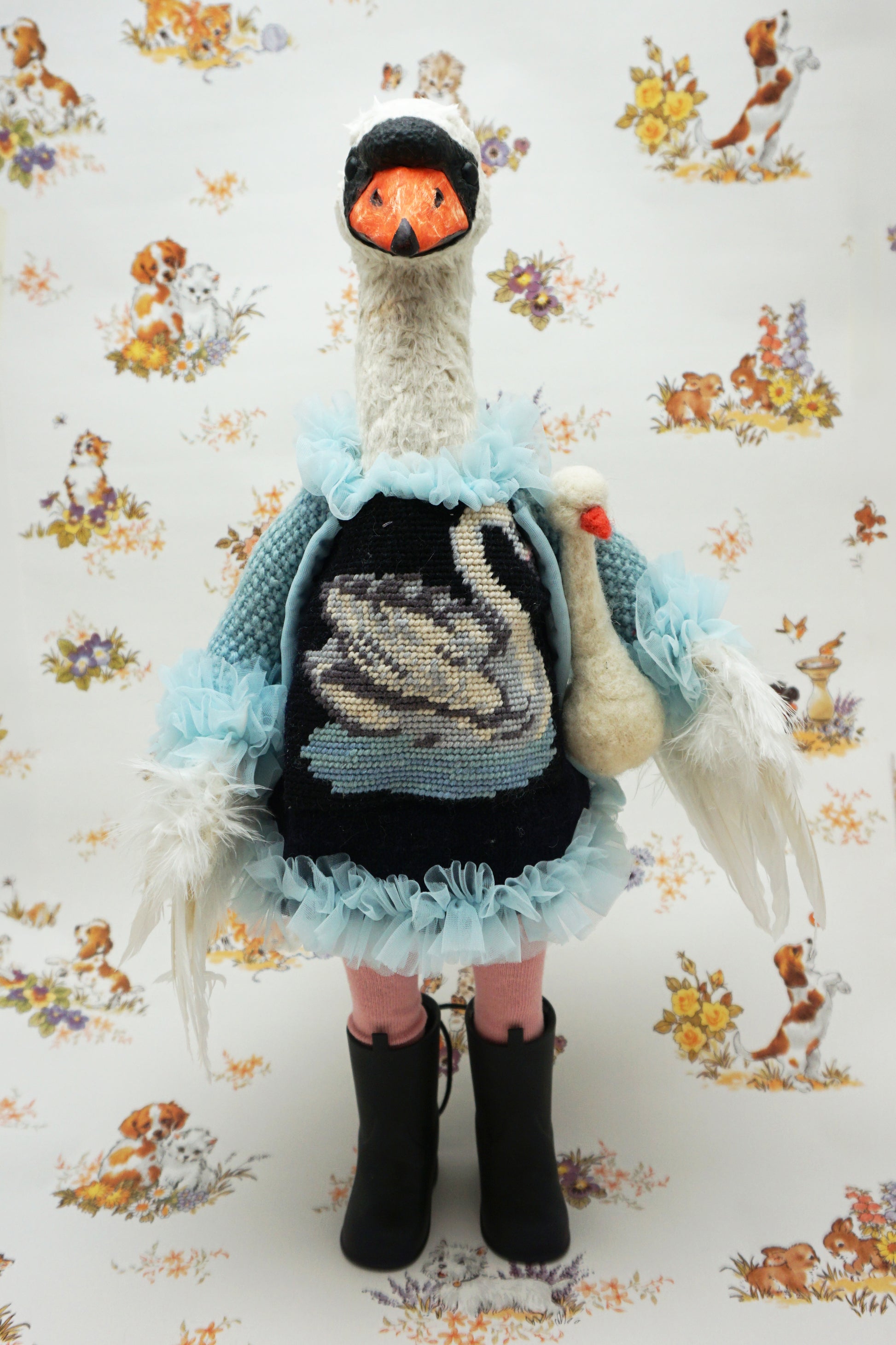 A plush swan holding a smaller swan toy dressed in a blue knitted jumper with a swan design, pink leggings, and black boots. It stands against a backdrop featuring illustrations of dogs and flowers. The swan's jumper has a fluffy blue collar and cuffs.