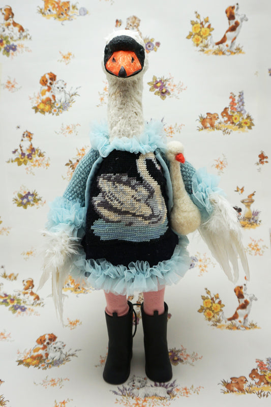 A plush swan holding a smaller swan toy dressed in a blue knitted jumper with a swan design, pink leggings, and black boots. It stands against a backdrop featuring illustrations of dogs and flowers. The swan's jumper has a fluffy blue collar and cuffs.