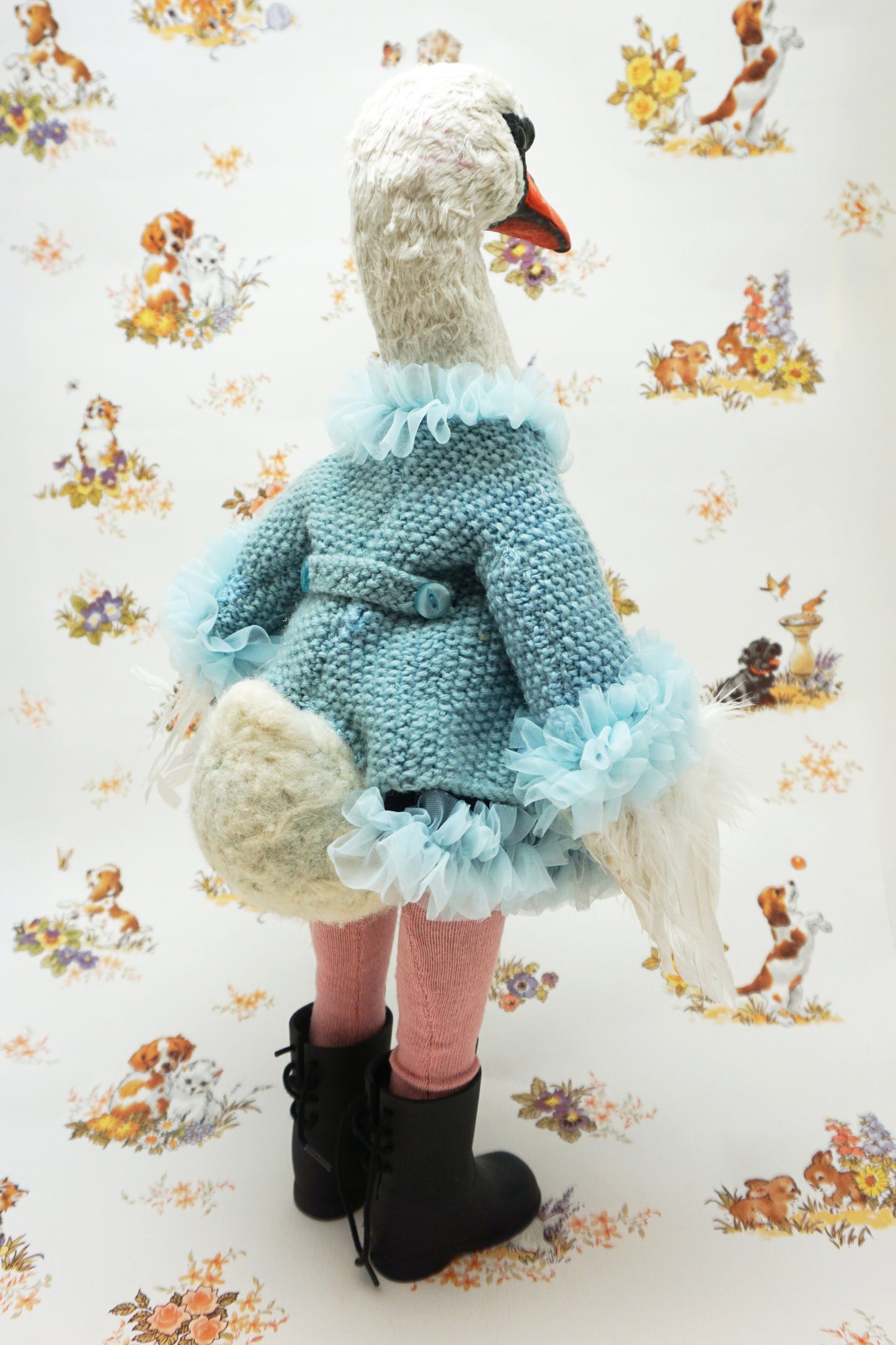 A plush swan holding a smaller swan toy dressed in a blue knitted jumper with a swan design, pink leggings, and black boots. It stands against a backdrop featuring illustrations of dogs and flowers. The swan's jumper has a fluffy blue collar and cuffs.
