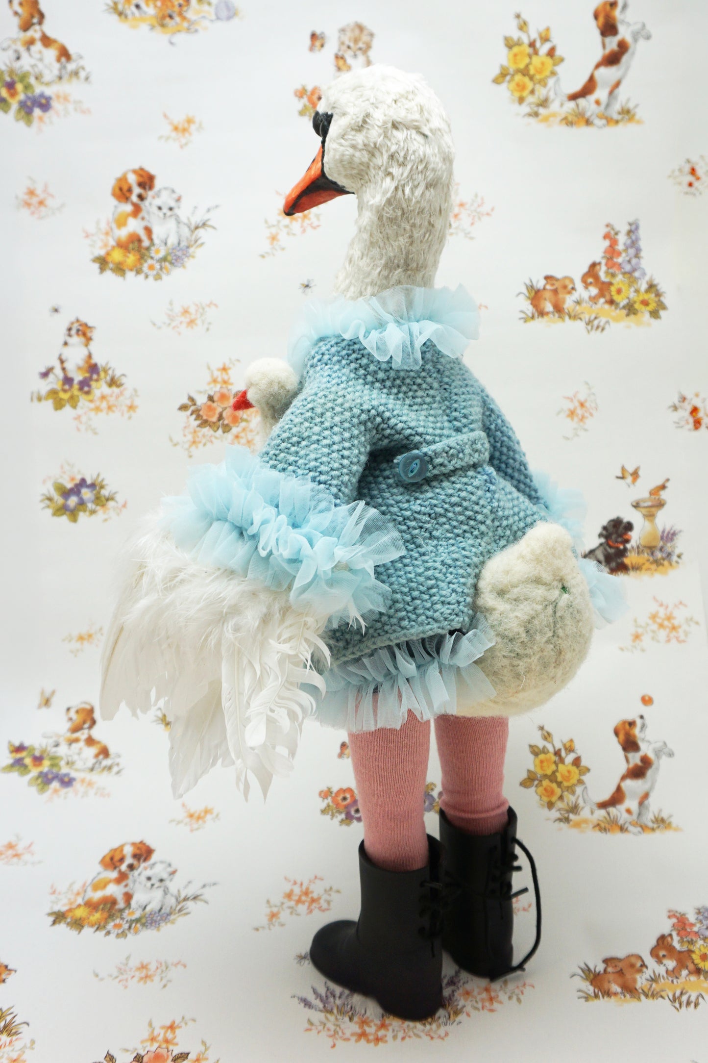 A plush swan holding a smaller swan toy dressed in a blue knitted jumper with a swan design, pink leggings, and black boots. It stands against a backdrop featuring illustrations of dogs and flowers. The swan's jumper has a fluffy blue collar and cuffs.