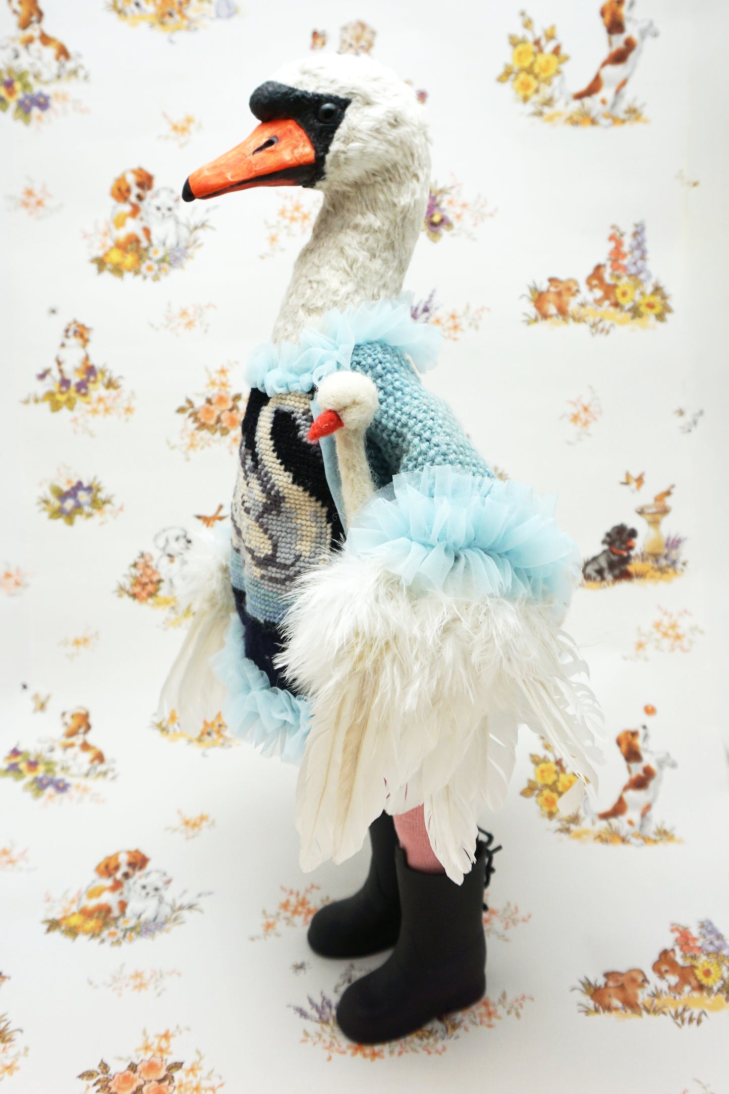 A plush swan holding a smaller swan toy dressed in a blue knitted jumper with a swan design, pink leggings, and black boots. It stands against a backdrop featuring illustrations of dogs and flowers. The swan's jumper has a fluffy blue collar and cuffs.