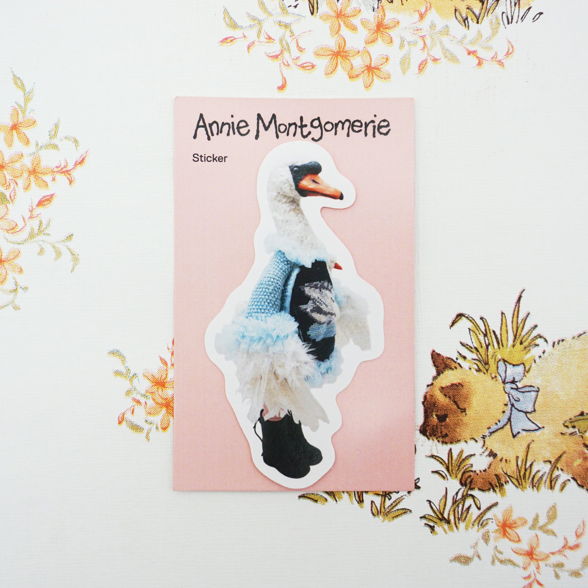 A die-cut sticker featuring a white swan wearing a blue knit sweater and black shoes, placed on a pink backing card labelled 'Annie Montgomerie Sticker' against a floral background.