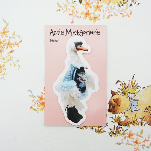 A die-cut sticker featuring a white swan wearing a blue knit sweater and black shoes, placed on a pink backing card labelled 'Annie Montgomerie Sticker' against a floral background.