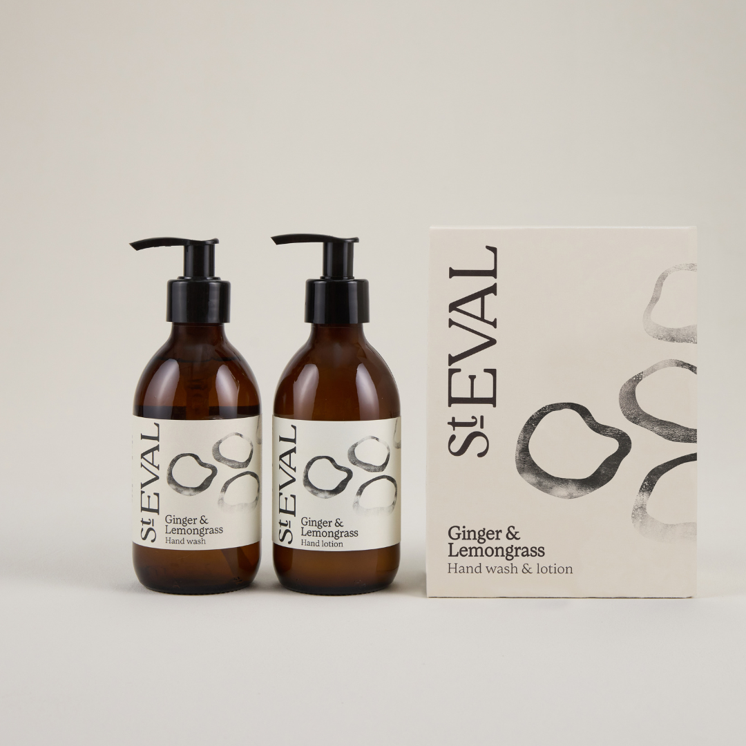 Two brown glass jars with pumps sitting next to a beige box with a foliage pattern on the right hand side on a plain grey background, featuring the branding 'St Eval' in black text.