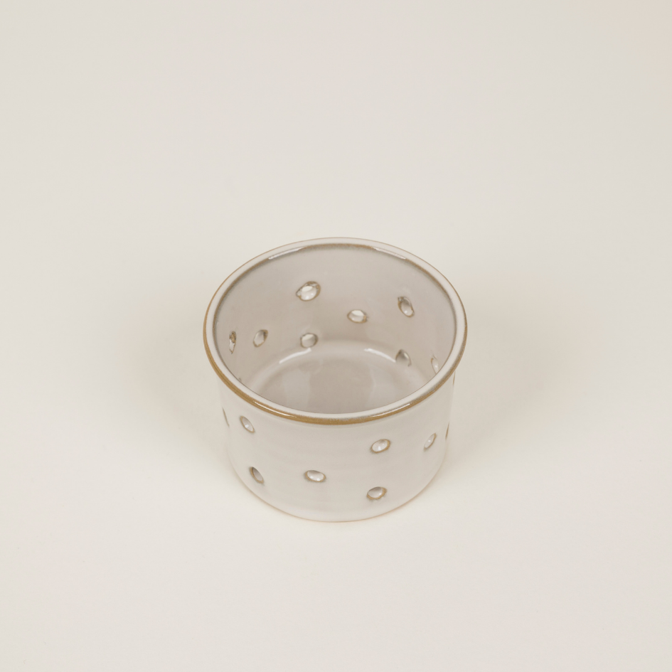 A small cylindrical ceramic candle holder with a glossy white glaze, featuring evenly spaced circular cut-outs around the body and a thin metallic gold rim, set against a plain off-white background.