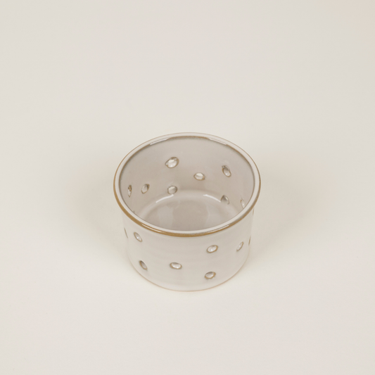 A small cylindrical ceramic candle holder with a glossy white glaze, featuring evenly spaced circular cut-outs around the body and a thin metallic gold rim, set against a plain off-white background.