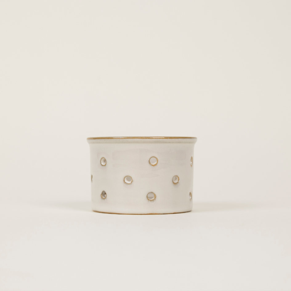 A small cylindrical ceramic tealight holder with a matte off-white finish, decorated with small metallic gold dots and a gold rim, set against a plain light background.