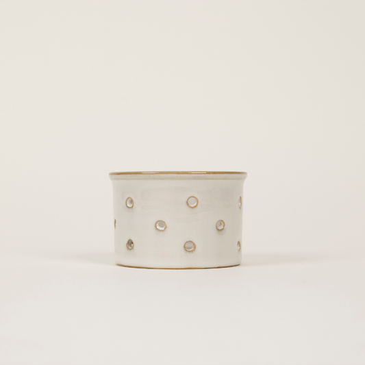 A small cylindrical ceramic tealight holder with a matte off-white finish, decorated with small metallic gold dots and a gold rim, set against a plain light background.