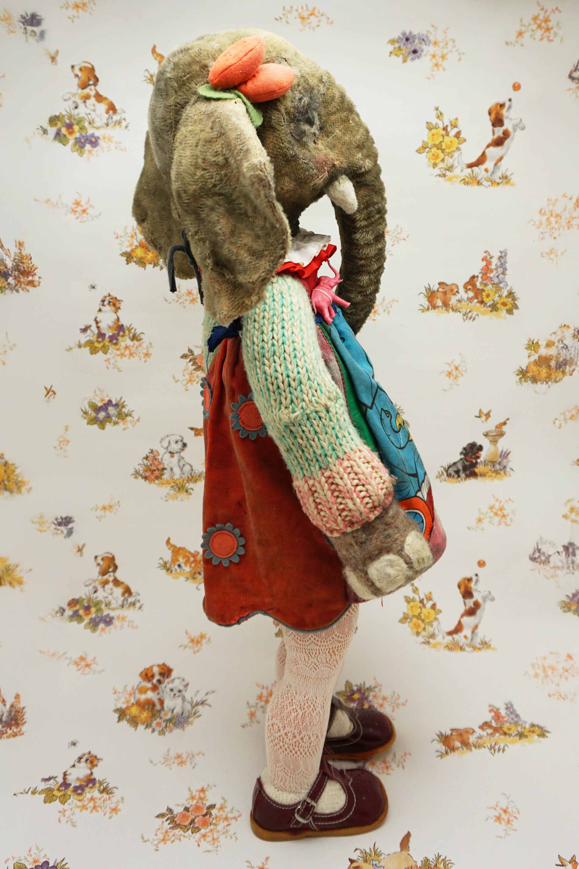 A plush elephant stands upright, wearing a colourful dress with a dog and fish design, a knitted green cardigan, pink tights, and red shoes. It has a pink bow on one ear. The background features whimsical animal illustrations.