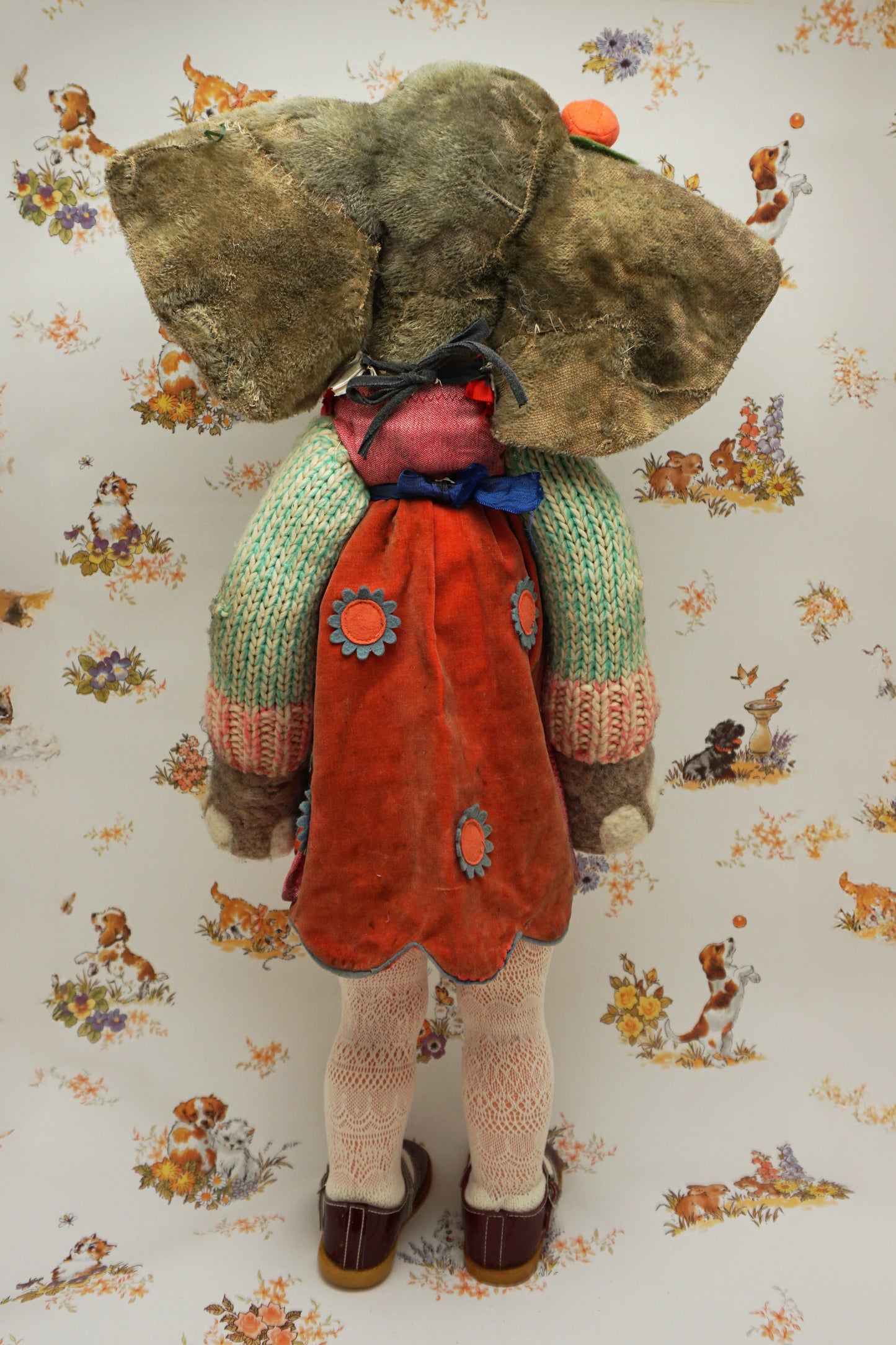 A plush elephant stands upright, wearing a colourful dress with a dog and fish design, a knitted green cardigan, pink tights, and red shoes. It has a pink bow on one ear. The background features whimsical animal illustrations.