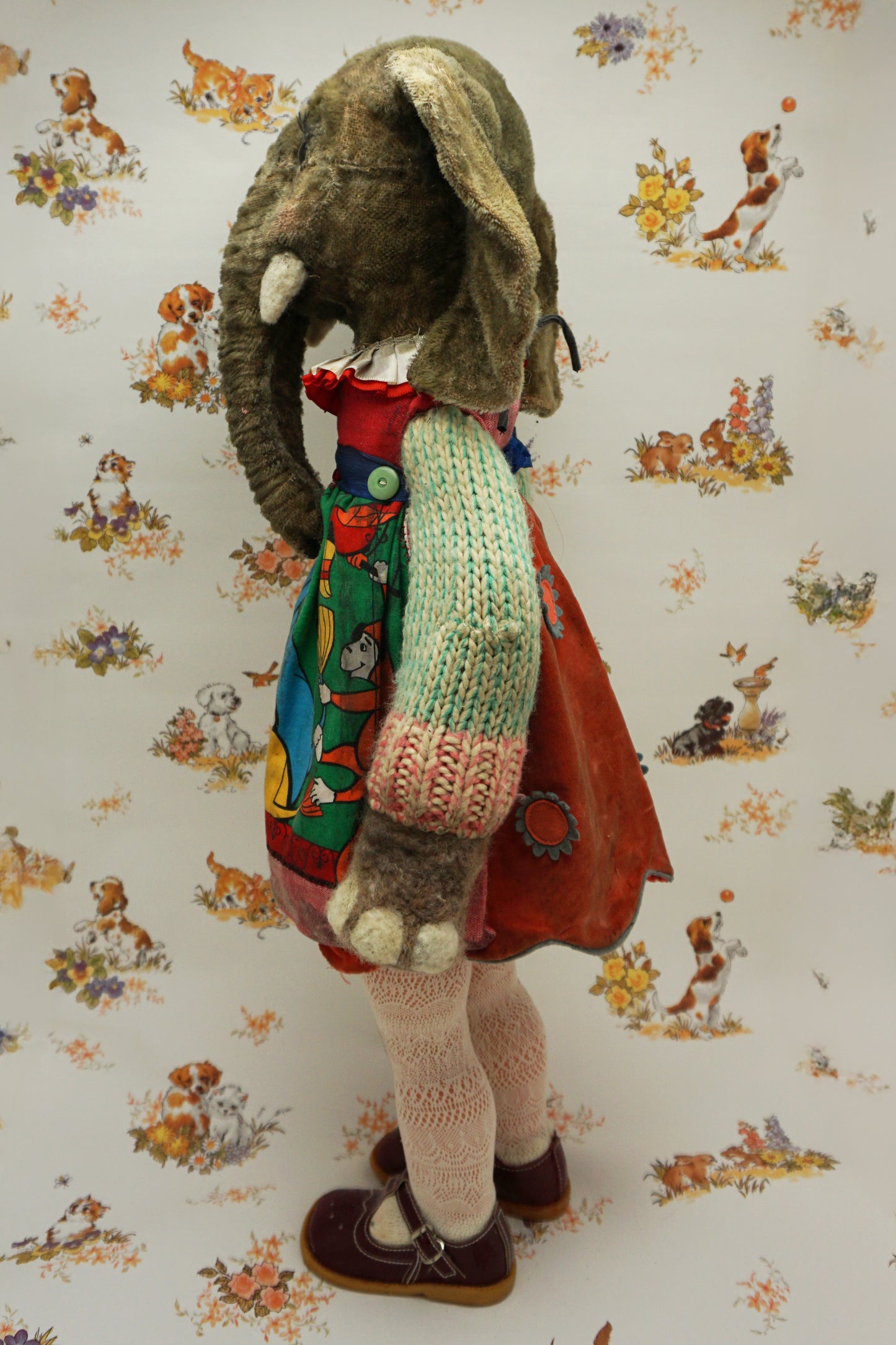 A plush elephant stands upright, wearing a colourful dress with a dog and fish design, a knitted green cardigan, pink tights, and red shoes. It has a pink bow on one ear. The background features whimsical animal illustrations.
