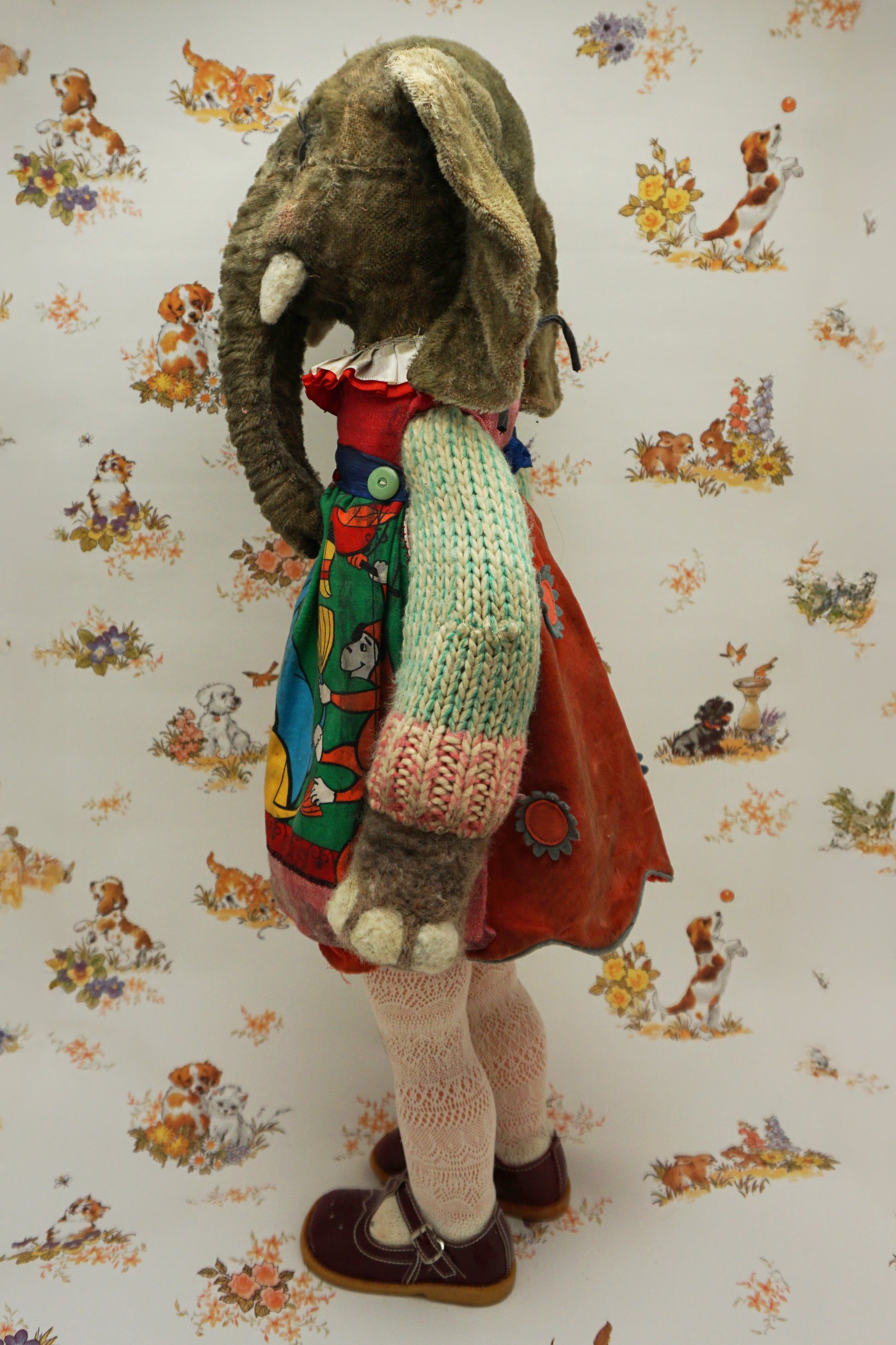 A plush elephant stands upright, wearing a colourful dress with a dog and fish design, a knitted green cardigan, pink tights, and red shoes. It has a pink bow on one ear. The background features whimsical animal illustrations.