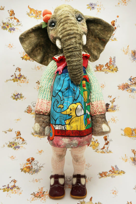 A plush elephant stands upright, wearing a colourful dress with a dog and fish design, a knitted green cardigan, pink tights, and red shoes. It has a pink bow on one ear. The background features whimsical animal illustrations.