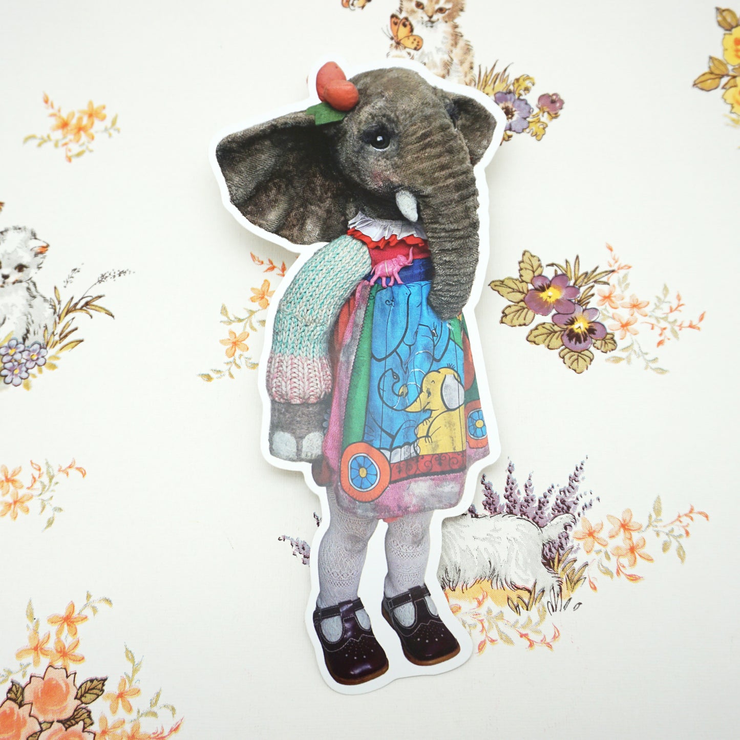 A die-cut postcard of an elephant wearing a colourful dress, knitted jumper, and black shoes. The elephant has a red bow on its head. The background features floral and animal patterns in soft pastel colours.