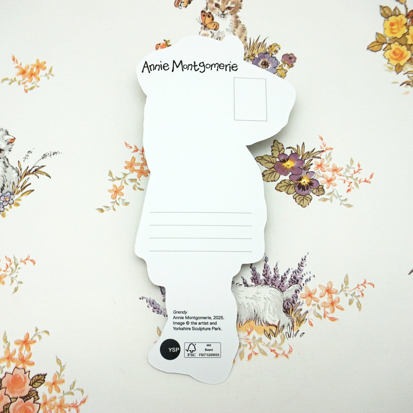 Silhouette-shaped postcard on floral wallpaper. The card features lines for writing and a printed name at the top. The background has illustrations of animals and flowers in soft colours.