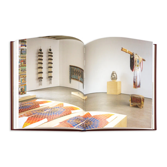 An open book showing a wide interior photograph of an art gallery or exhibition space, with colourful textile and wall-hanging artworks displayed in a room with white walls and a concrete floor.