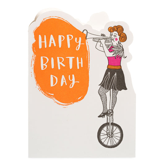 Greetings card with a design of a woman on a unicycle playing a trumpet, with 'Happy Birthday' text on an orange background.