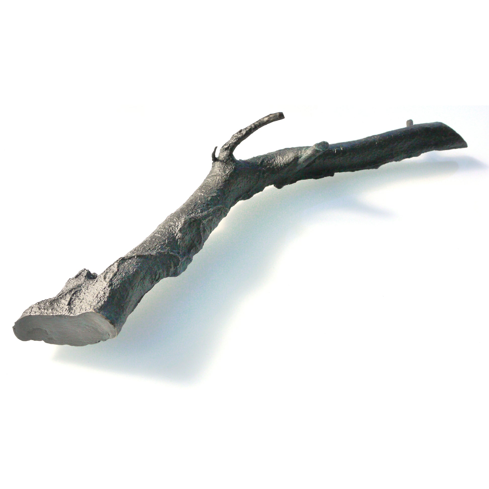 A textured, dark metallic branch-like sculpture lies on a light background. The piece features naturalistic details, including knots and curves, resembling a real twig, but is crafted from a solid material, giving it an artistic and modern appearance.